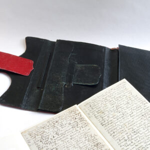 Das Bild zeigt eine schwarz-rote Brieftasche Richard Wagners auf weißem Untergrund, davor ein Notizbuch mit handschriftlichen Tagebucheinträgen, die der Komponist im Alter von 22 Jahren begann und die später zur Grundlage seiner Autobiographie wurden.